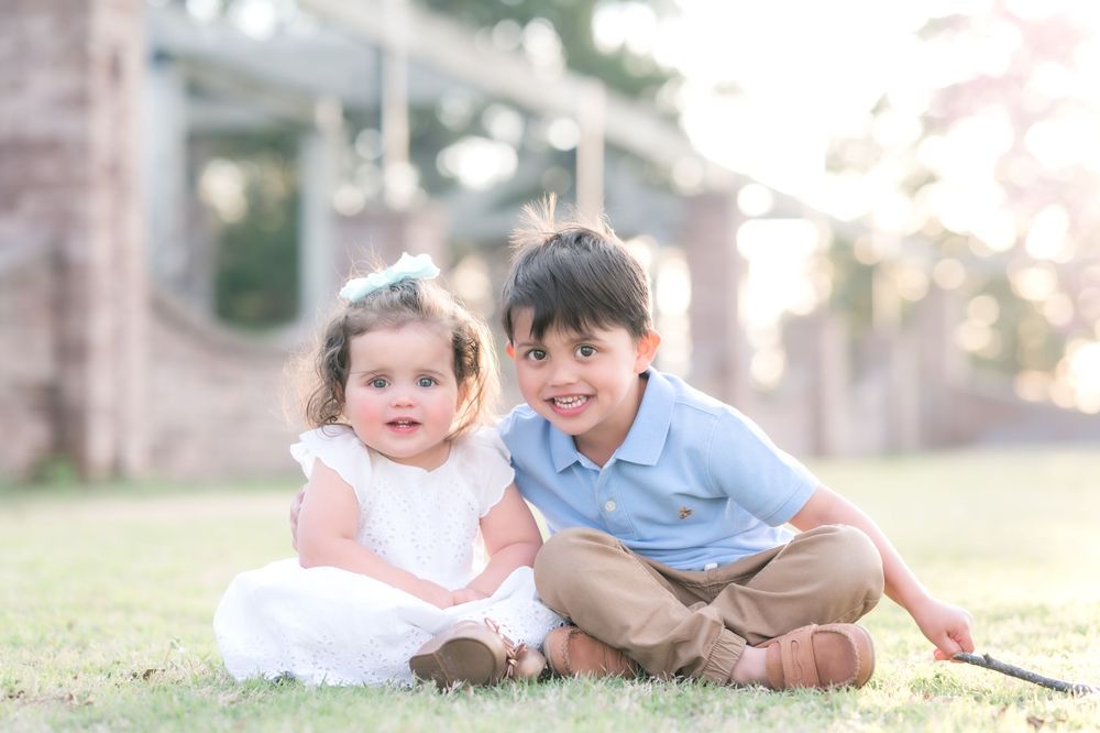 Two young children sit together smiling on grass wearing light colored clothing in a sunny outdoor setting.