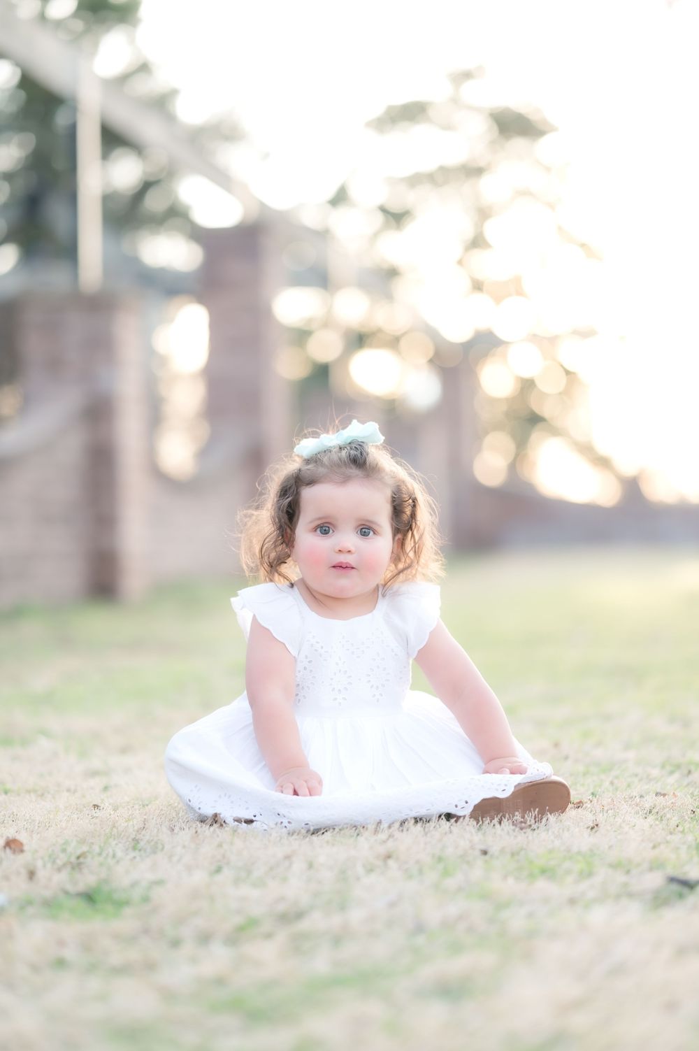 A small child in a white lacy dress sits on grass during golden hour with a dreamy bokeh background.