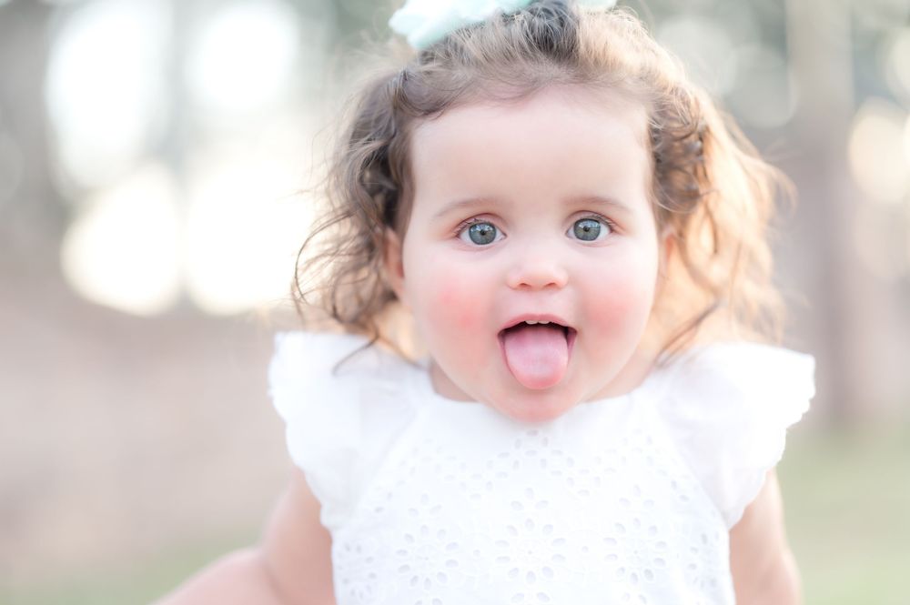 Sweet little toddler in white dress playfully sticks out tongue while being photographed outdoors in soft lighting.