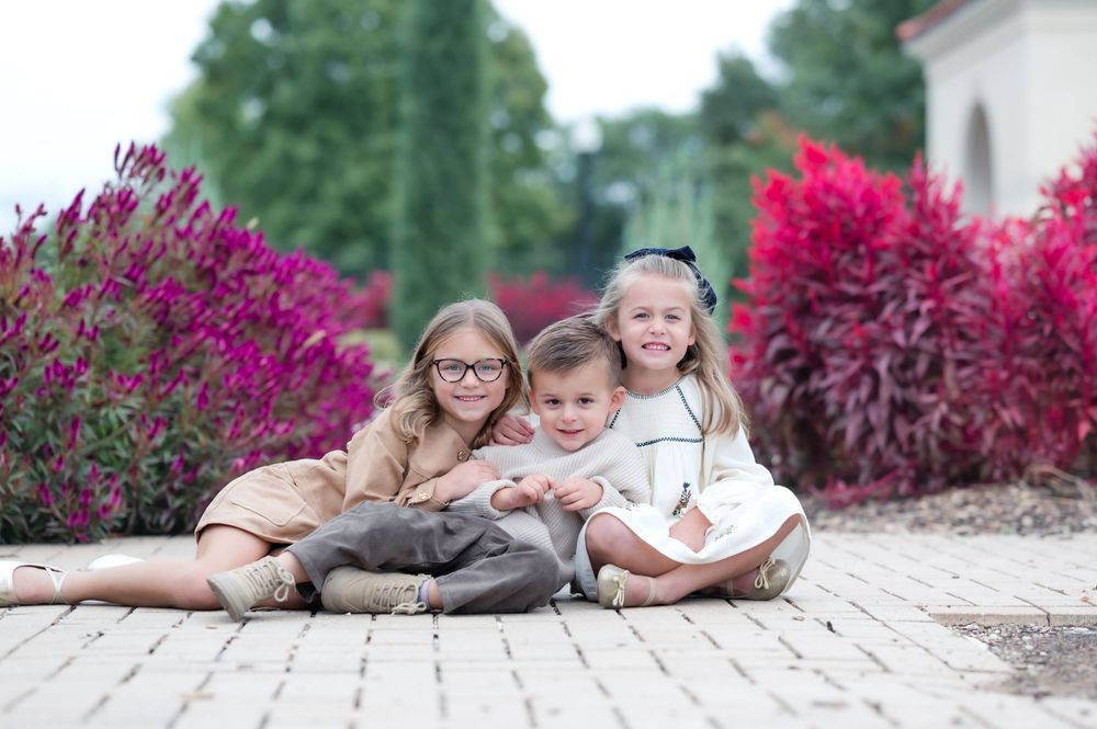 Children sitting together on a brick paver walkway with vibrant purple and red flowers in the background.