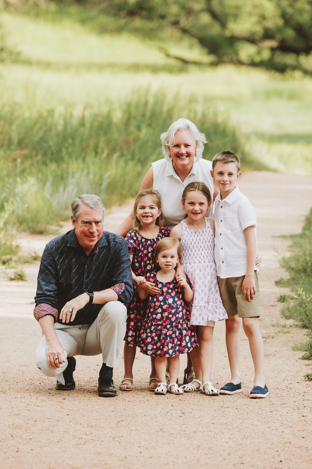 A family group poses together on a dirt path surrounded by green grass and trees on a sunny day.