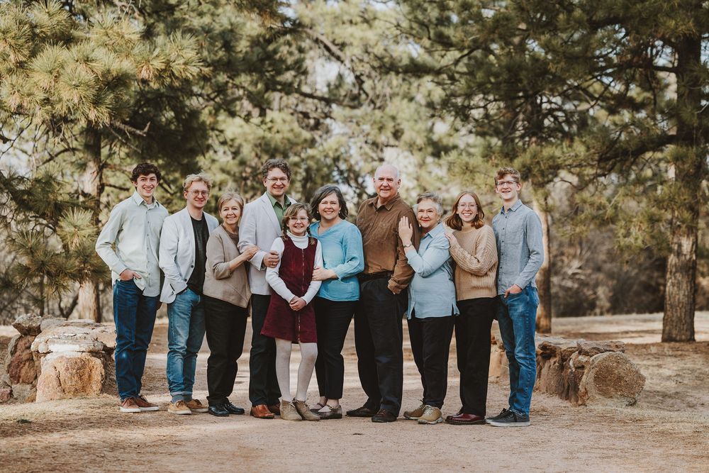Group of people wearing coordinated casual outfits posing together outdoors among pine trees on a sunny day.