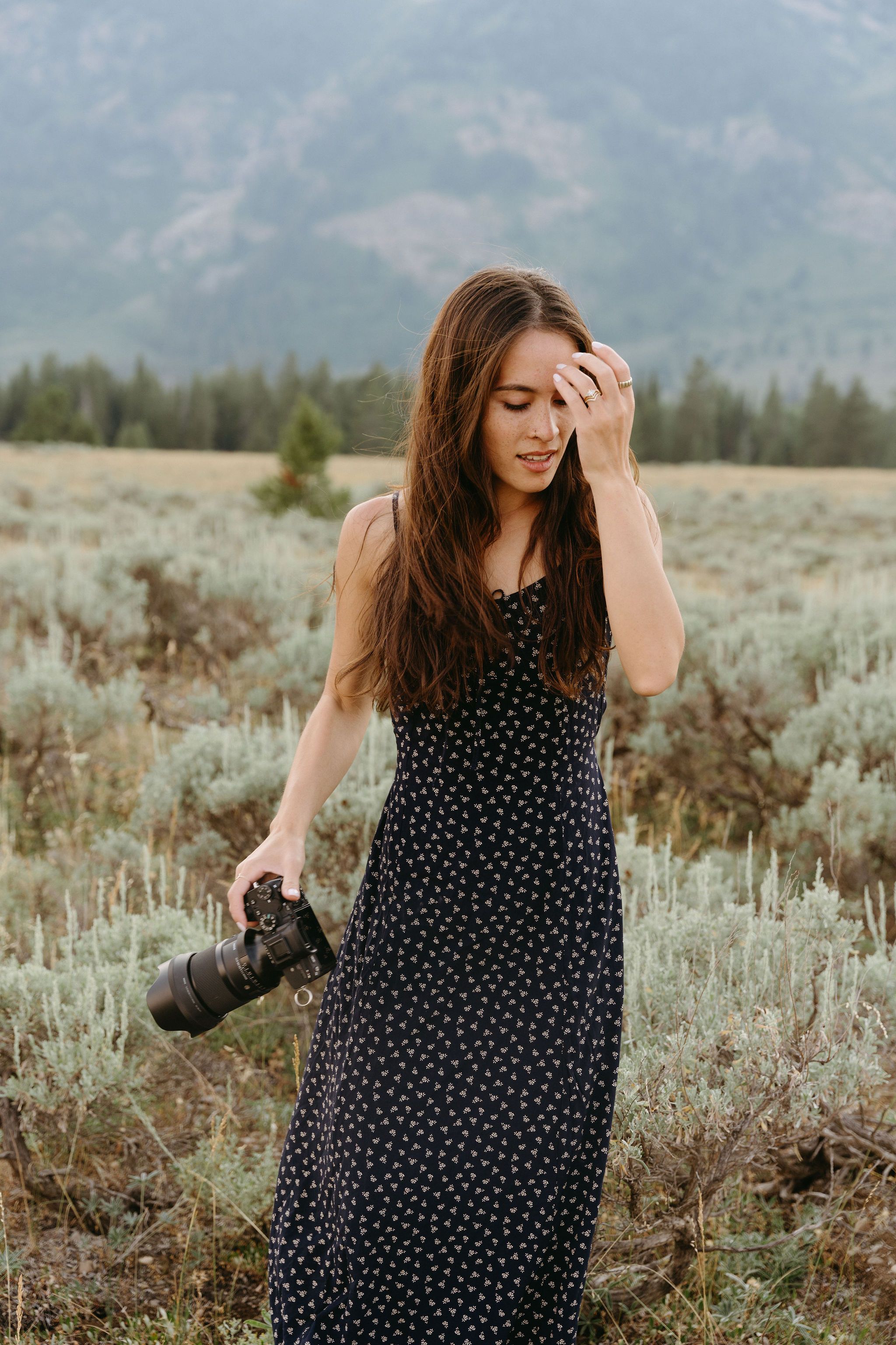 Person in polka dot maxi dress holds camera while standing in field with mountains in background.