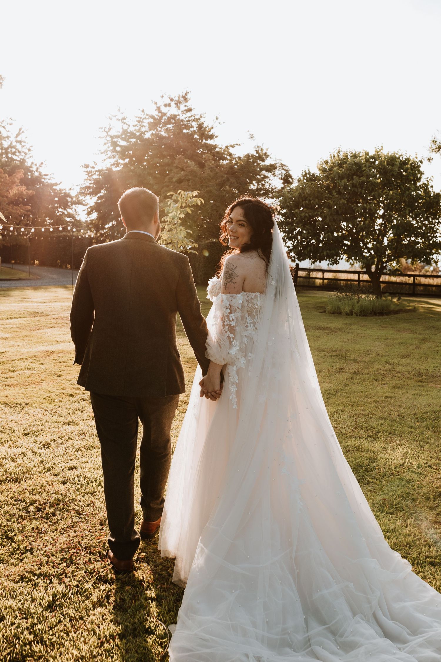 A wedding couple walks together across a sunlit grassy field during golden hour.