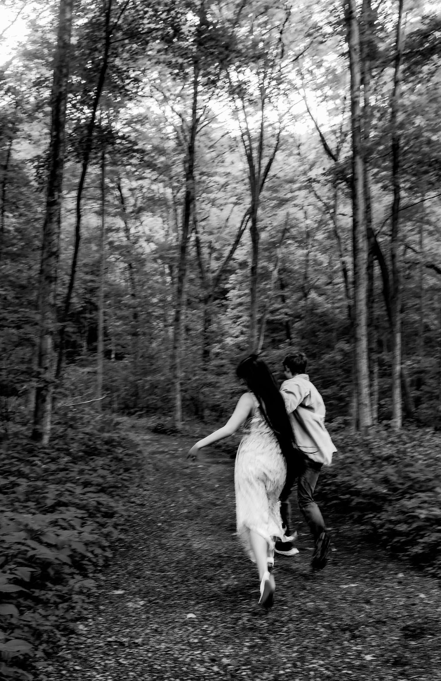 A black and white photograph of two figures running playfully down a wooded forest path.