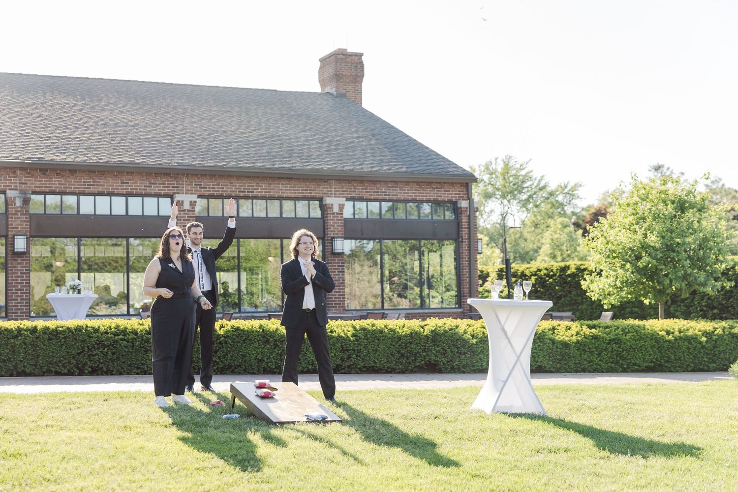 People standing on a manicured lawn in front of a rustic lodge building with hedges and a cocktail table.
