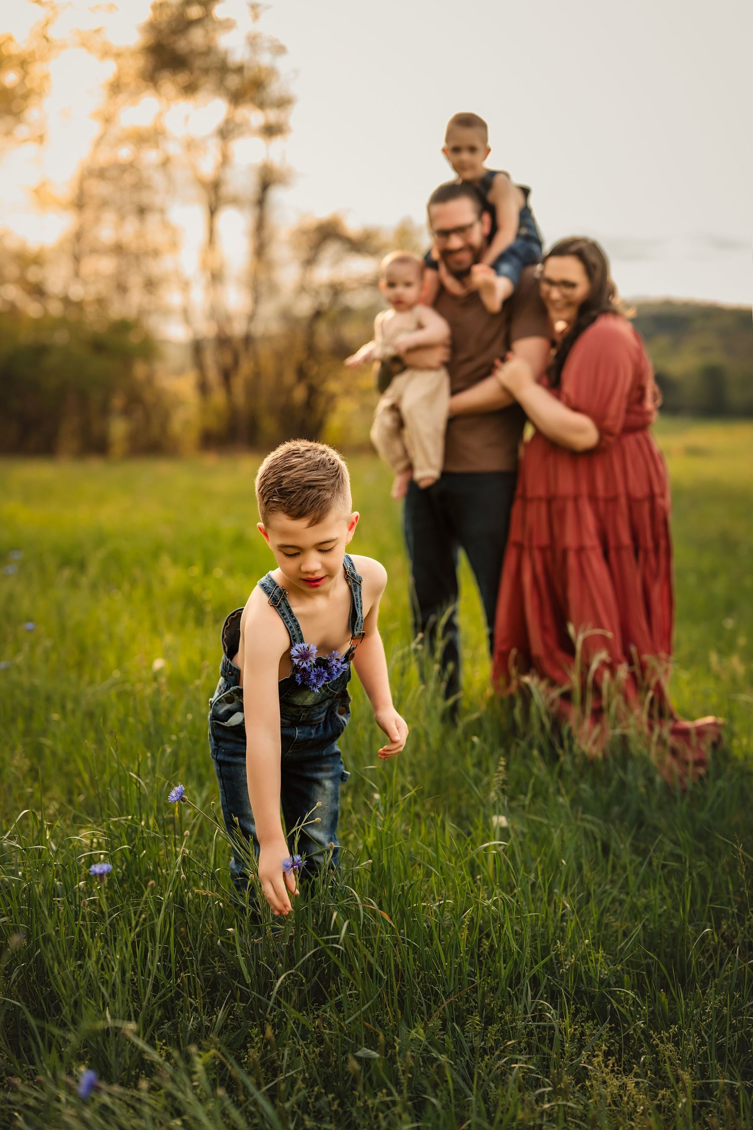 A boy in overalls explores a grassy meadow at sunset while a family watches from behind in a pastoral setting.