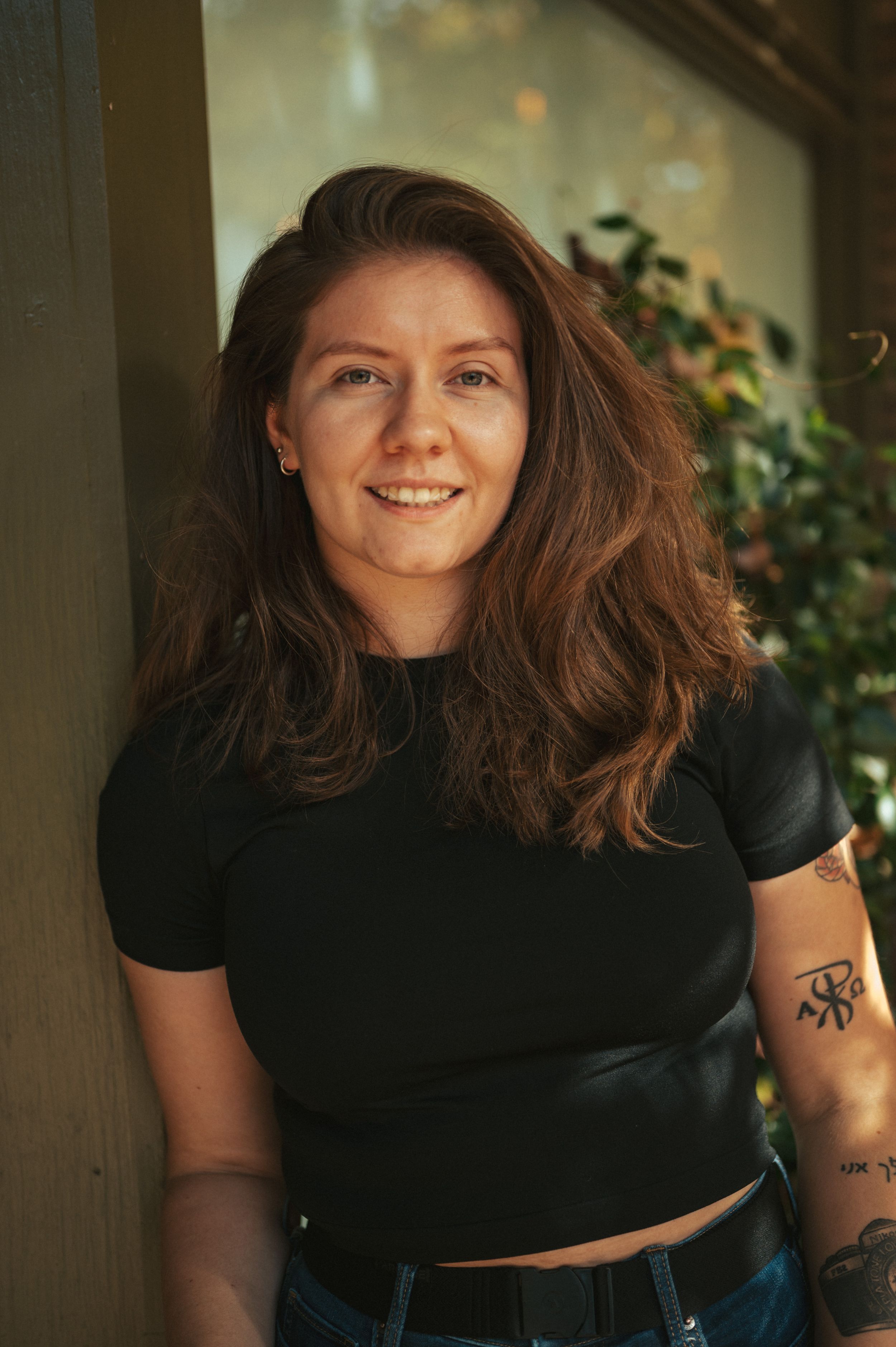 A warm portrait shows a smiling person with flowing brown hair wearing a black t-shirt against a sunlit background.