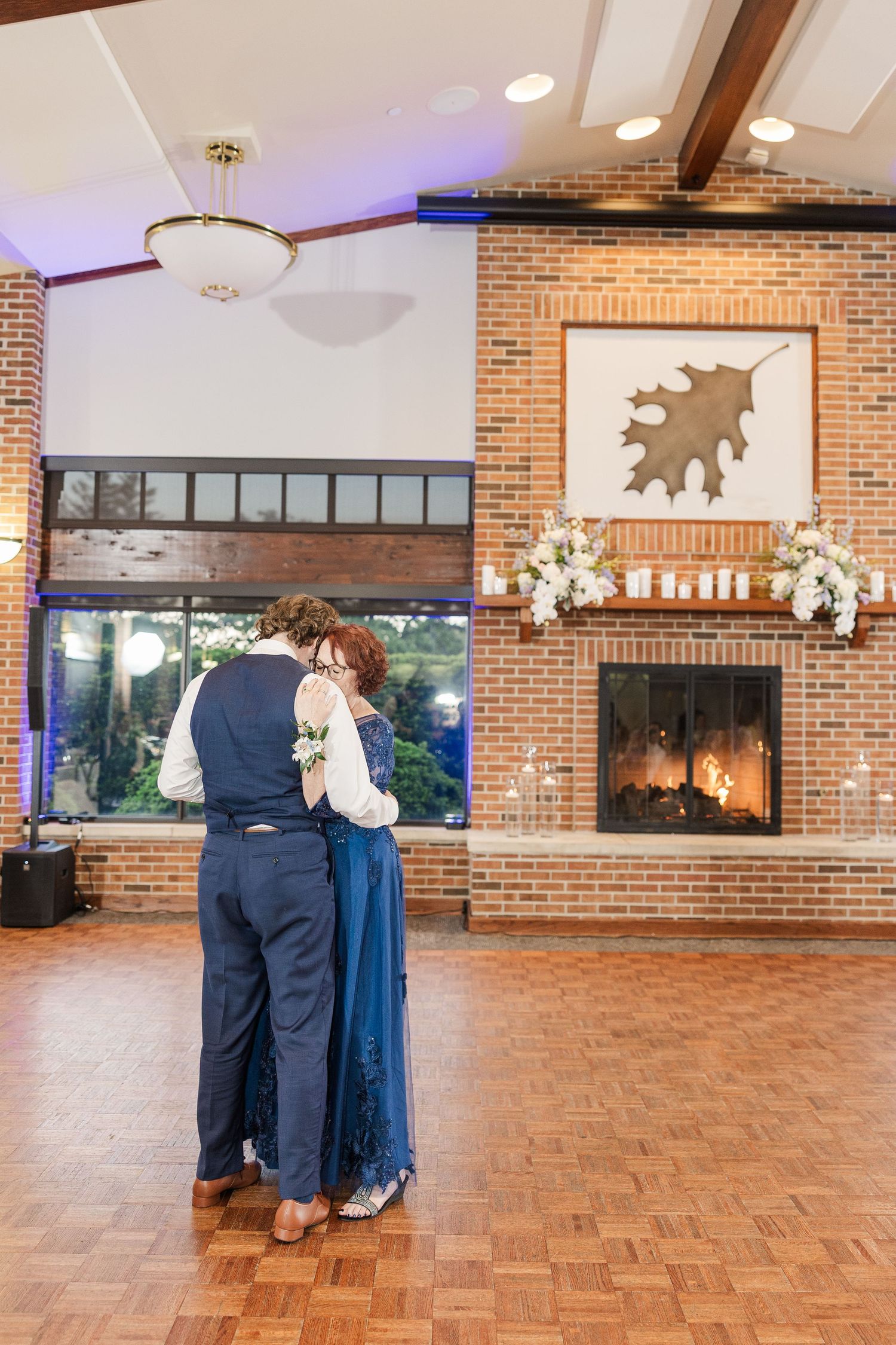 A couple shares a romantic dance on a parquet floor in front of a brick fireplace and chandelier-lit room.