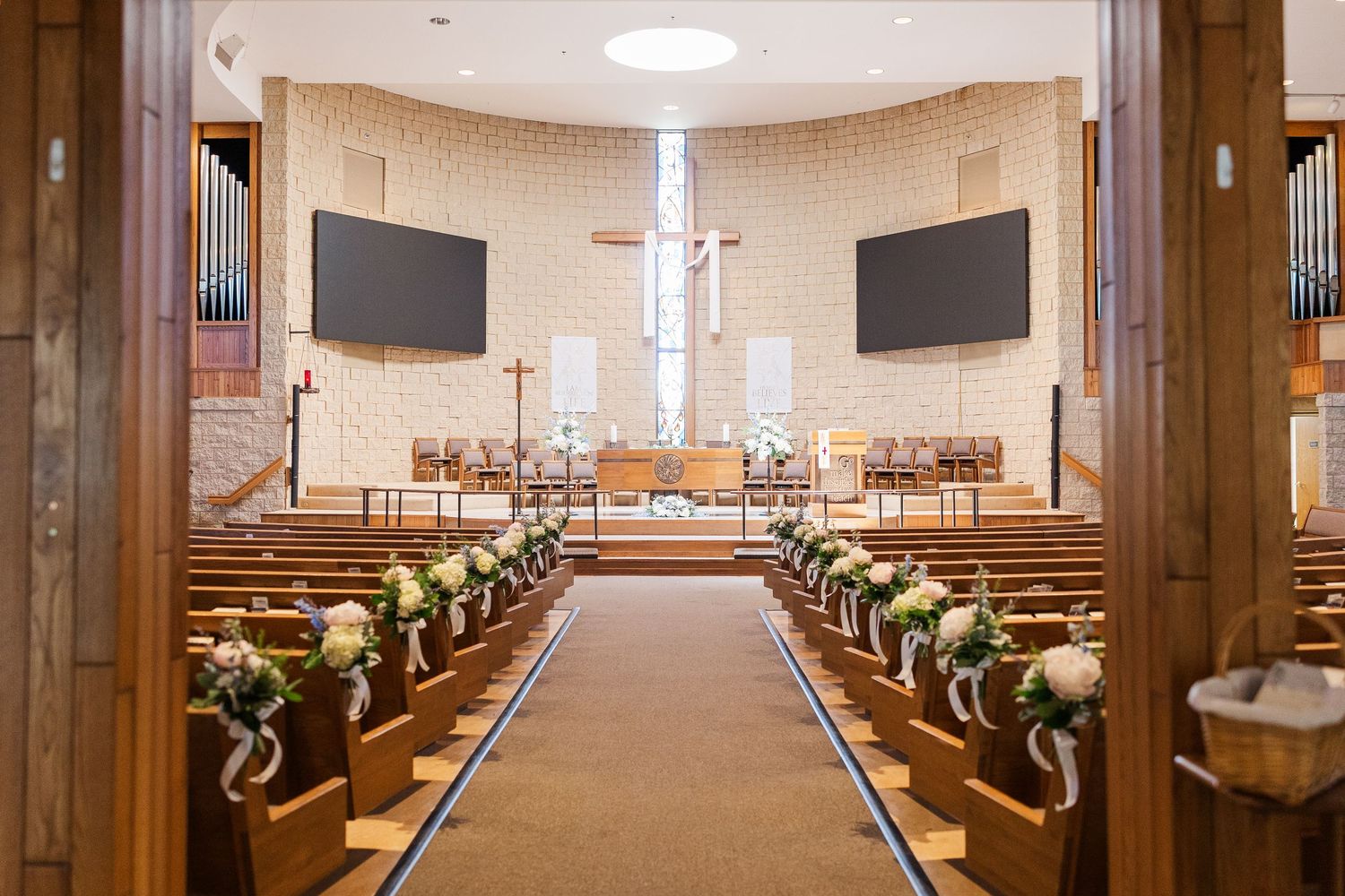 Modern church sanctuary interior with wooden pews, carpeted aisle, and dual video screens mounted on beige walls.