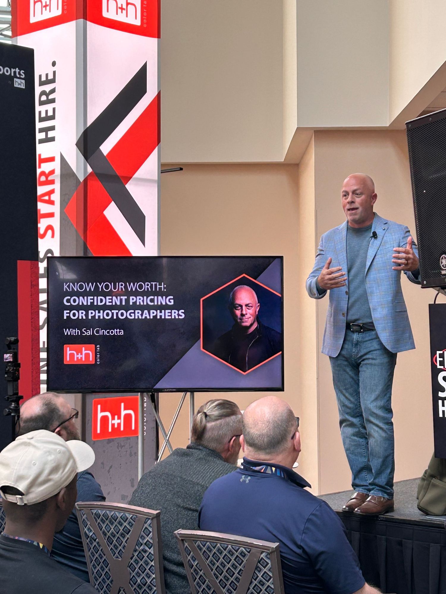 A speaker presents on stage at a photography conference with red and black branding displays and presentation screens.