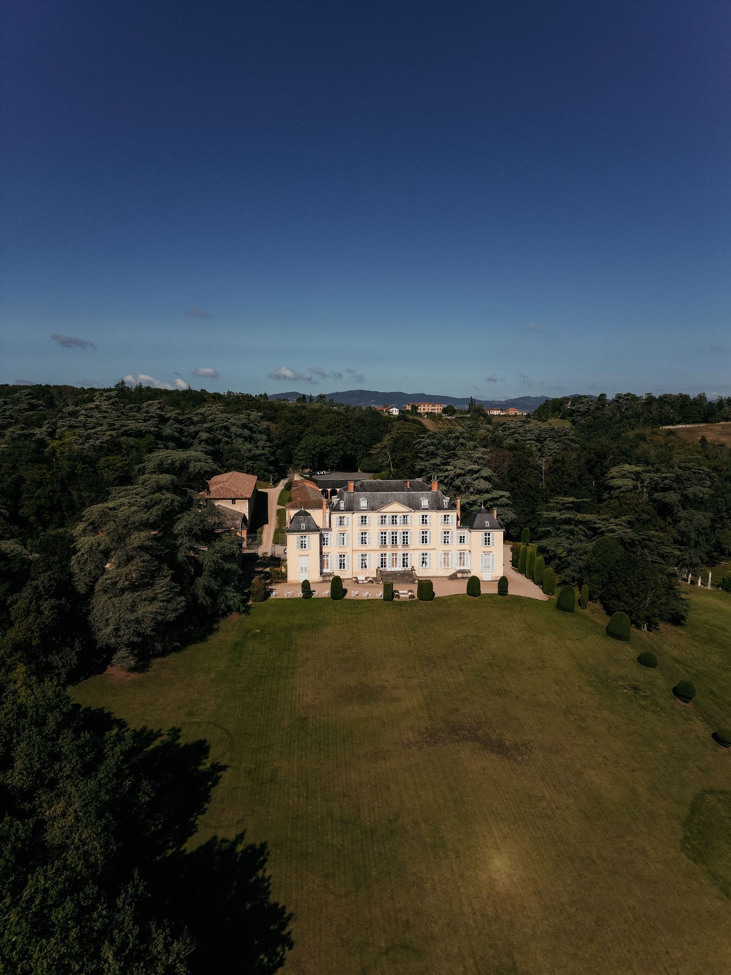 Aerial view of a grand white chateau mansion surrounded by lush green forest and manicured lawn on a sunny day.