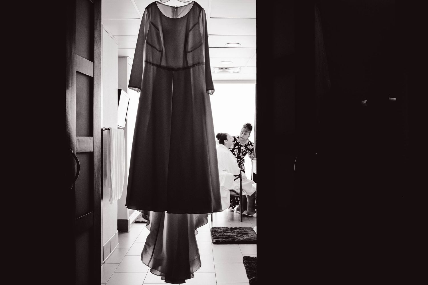 A black and white photograph of a long dark dress hanging in a doorway, casting dramatic shadows.