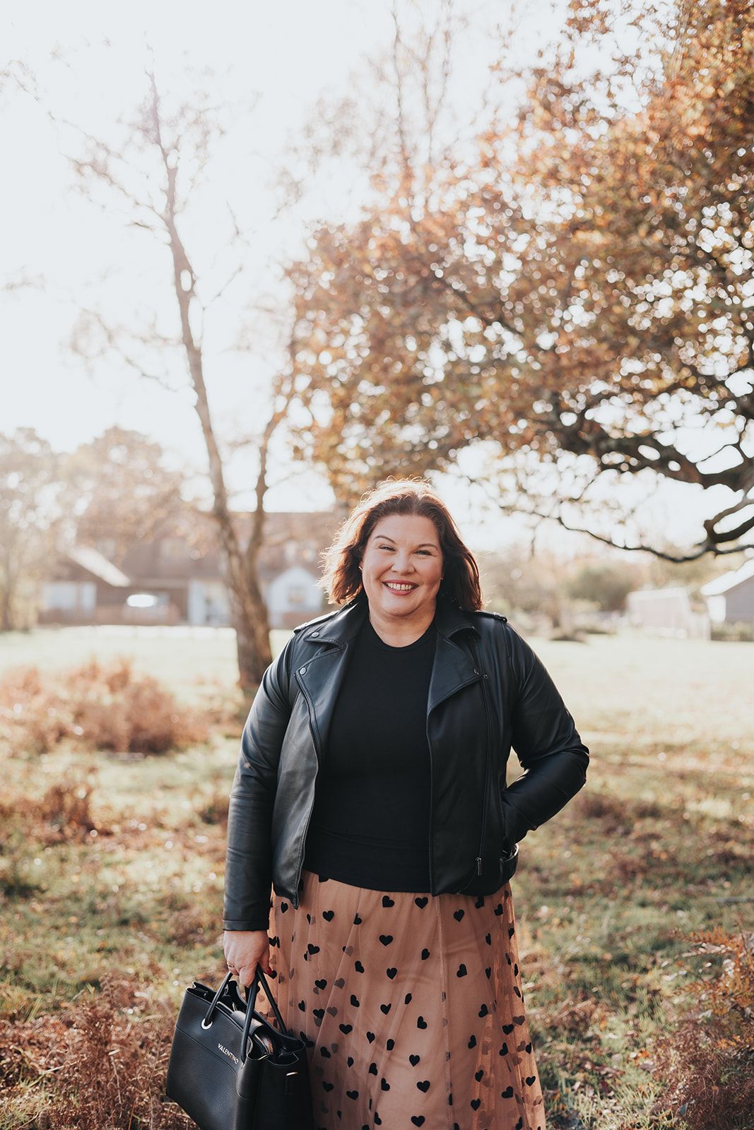 Someone poses outdoors in autumn wearing a black leather jacket and heart-patterned skirt while holding a handbag.
