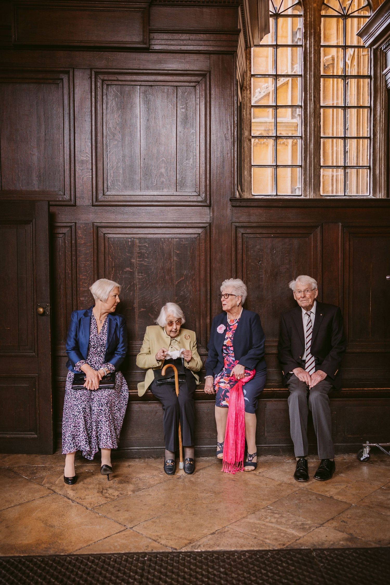 Four elderly people sitting together on a wooden bench in an elegant interior with paneled walls and vintage windows.