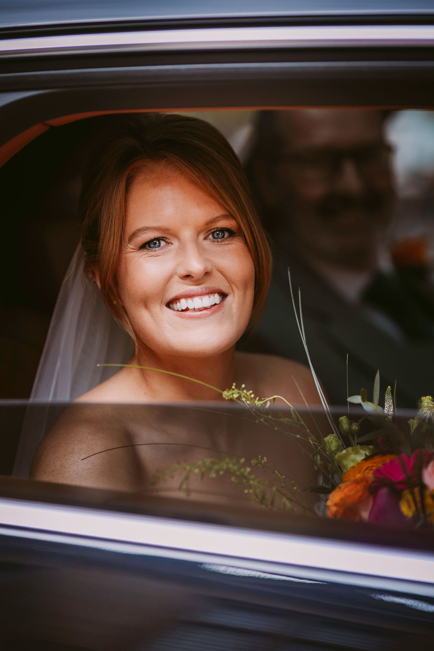 Smiling bride in a car looking through window with flowers and greenery visible.