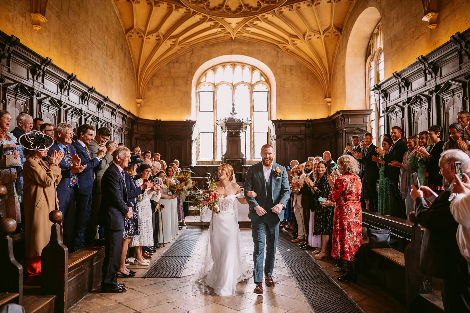A joyful couple walks down the aisle in a grand chapel with vaulted ceilings while guests stand in wooden pews.