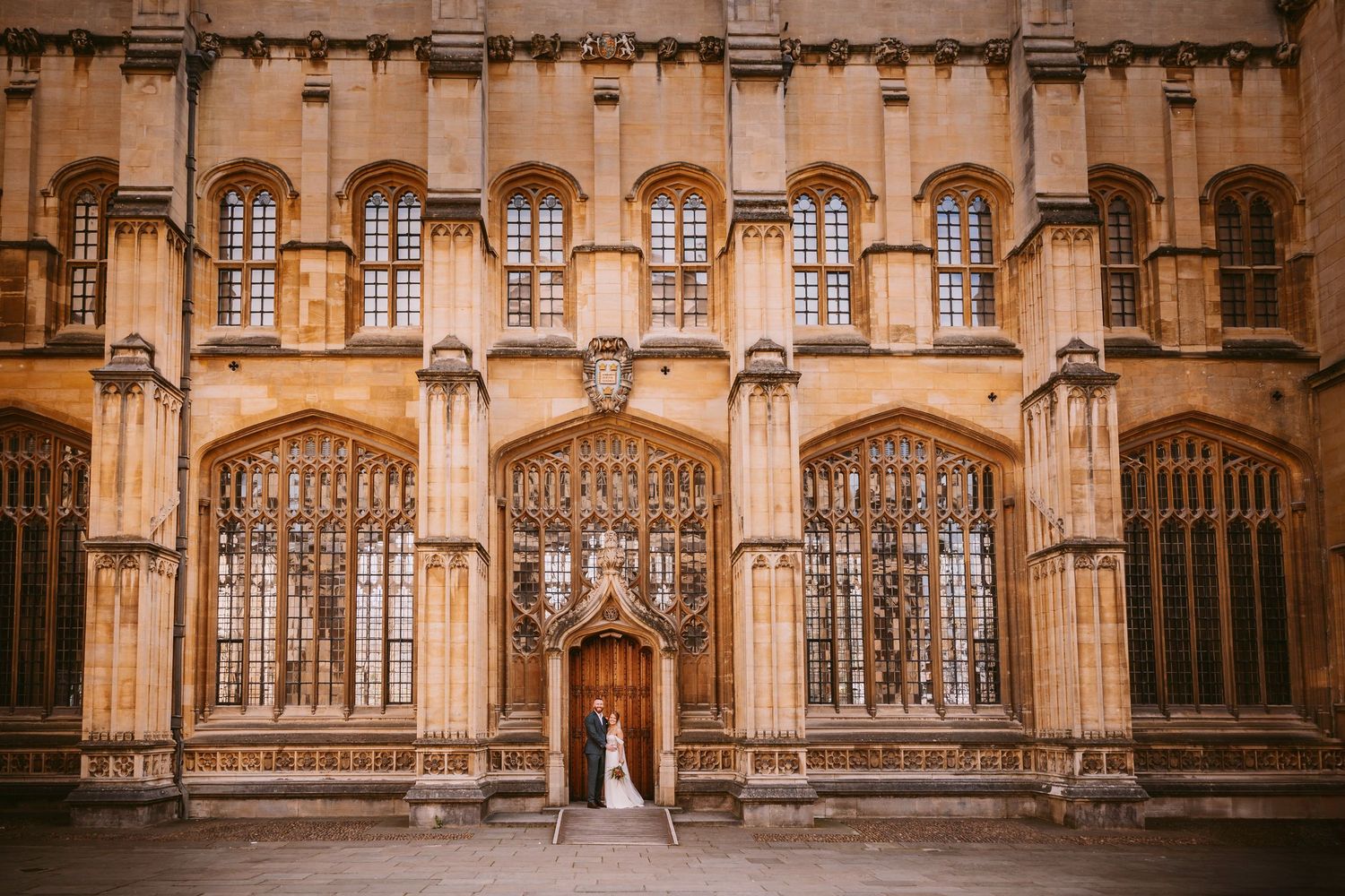 Historic Gothic college building with ornate stonework and windows frames a bride in a doorway.