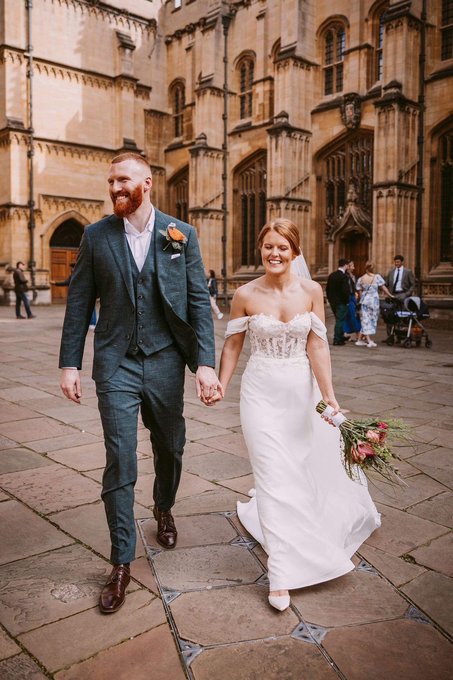 A newlywed couple walks together outside a Gothic cathedral with stone walls and arches.