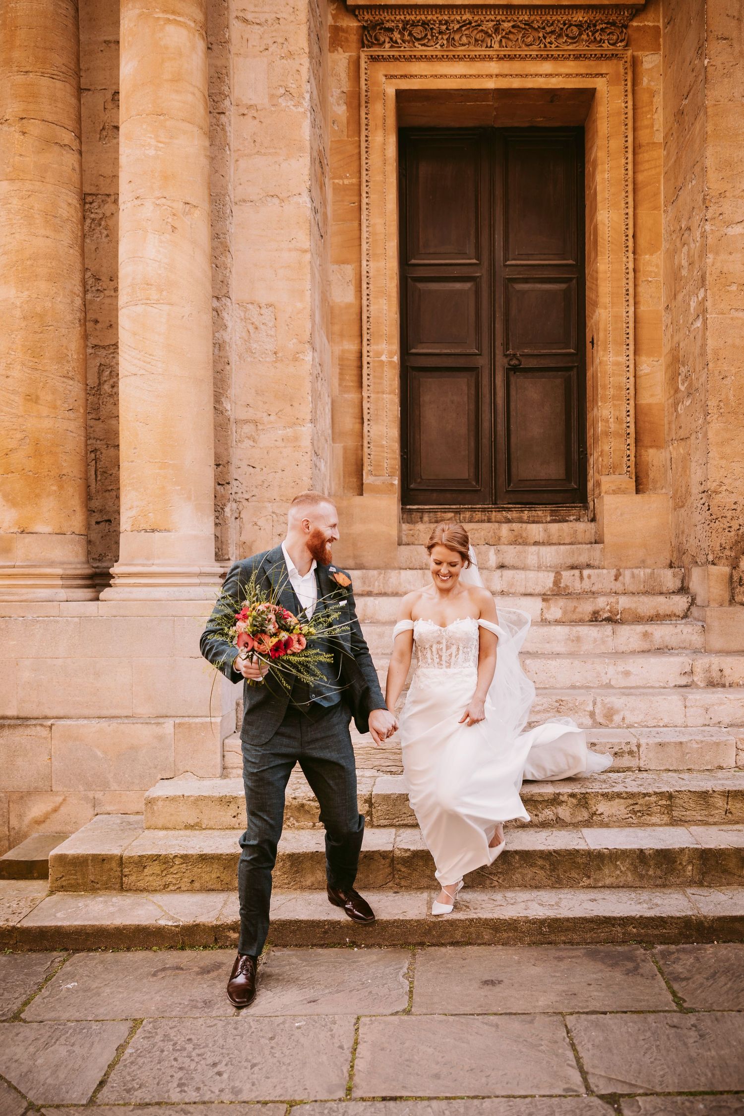 Wedding couple sitting casually on stone steps of a historic building with beige columns.