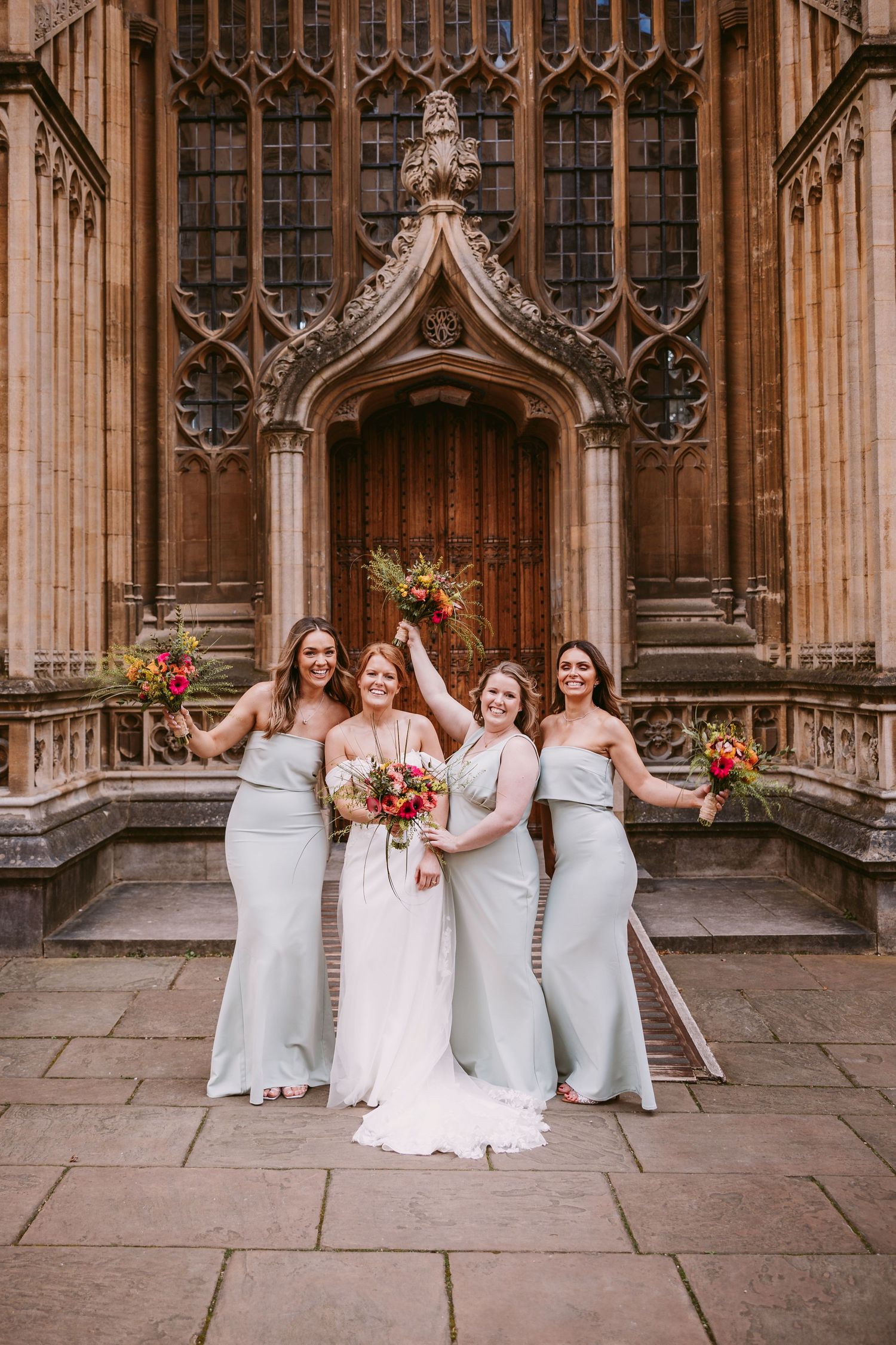 A bridal party poses in front of an ornate Gothic church entrance with stone archways and colorful bouquets.