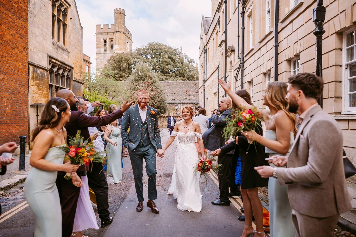 Wedding guests shower newlyweds with confetti on cobblestone street with historic church tower in background.