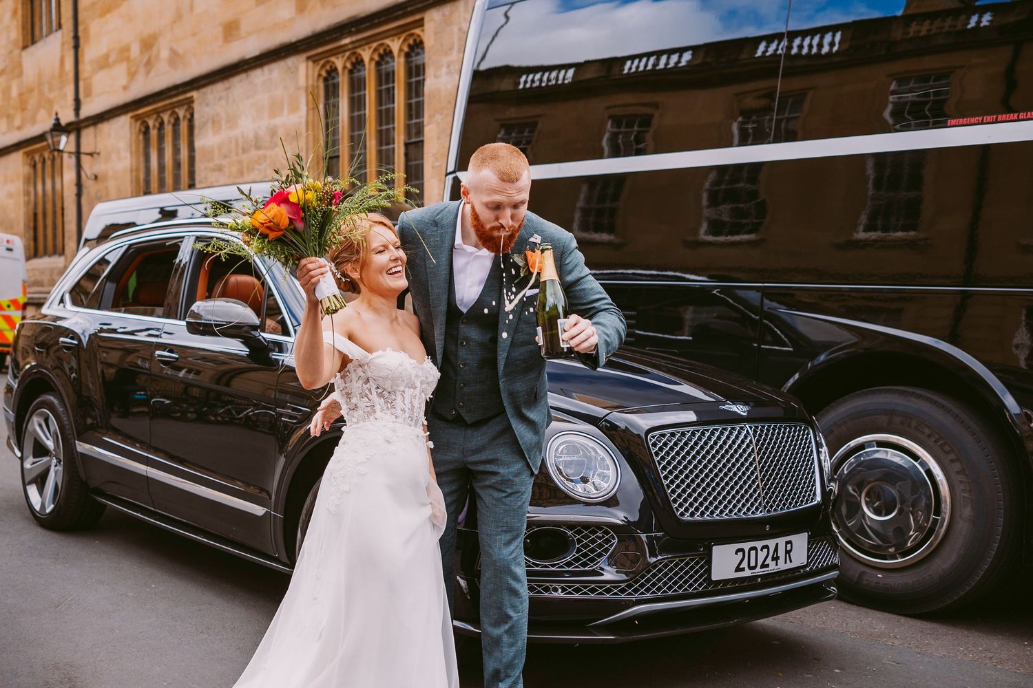 A couple in wedding attire walk together near a black luxury car on an urban street.