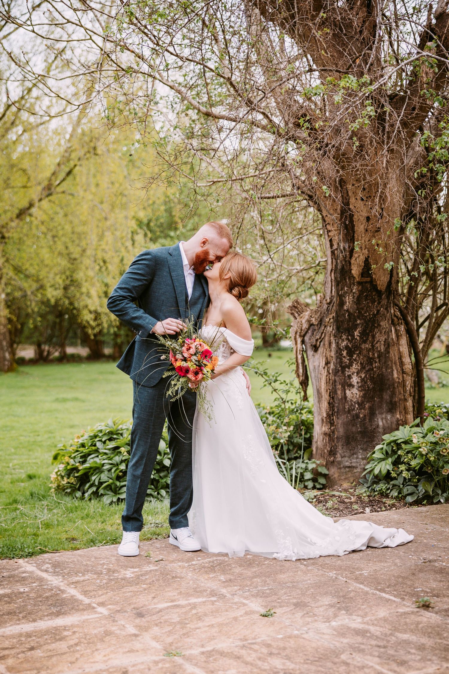 A wedding couple shares a romantic moment near a large tree in a garden setting.