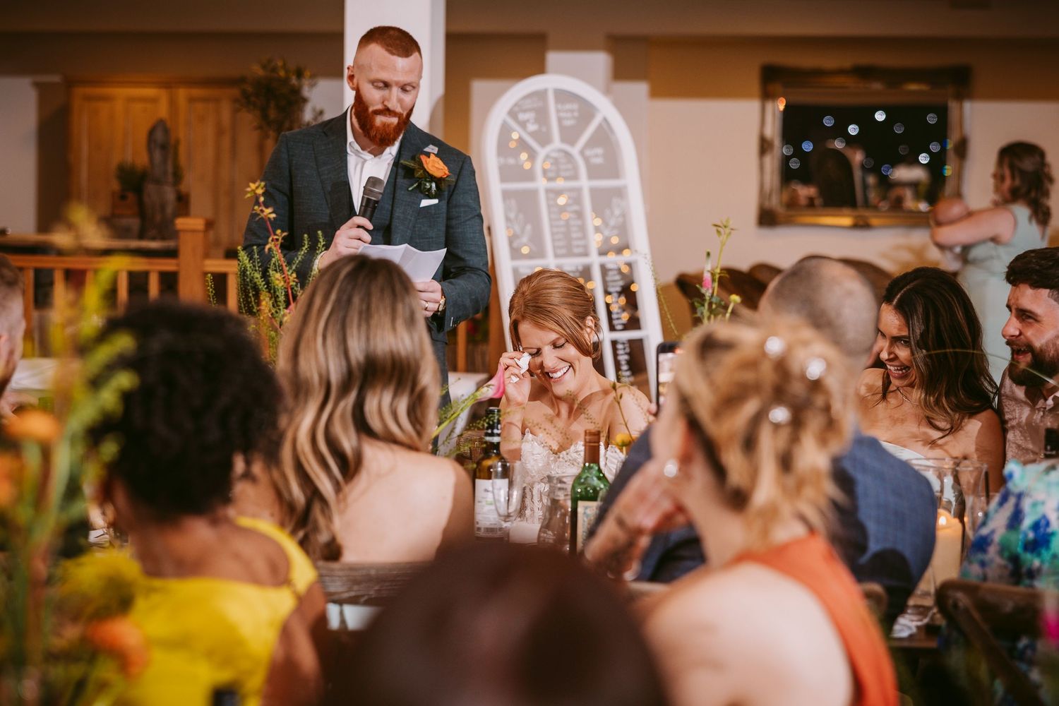 Wedding guests gathered around a table at a reception venue while someone delivers a speech.
