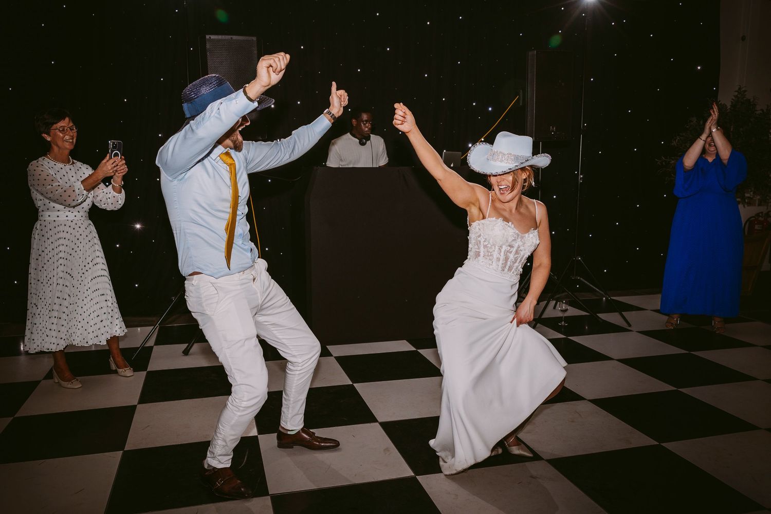 Wedding reception dancers in sync on a checkerboard floor with mood lighting and scattered onlookers.