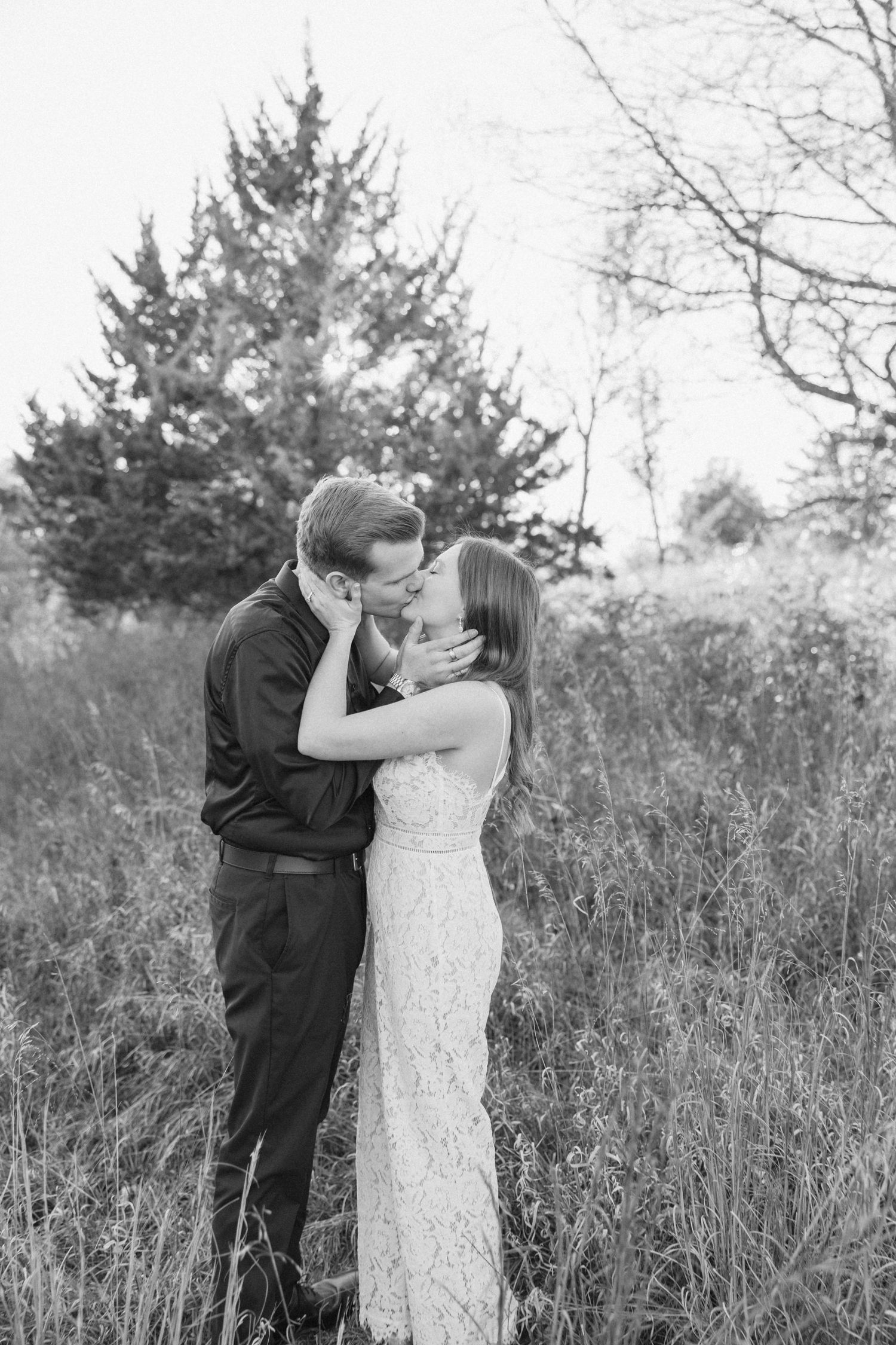 A couple embraces in a romantic moment outdoors with evergreen trees and tall grass in the background.