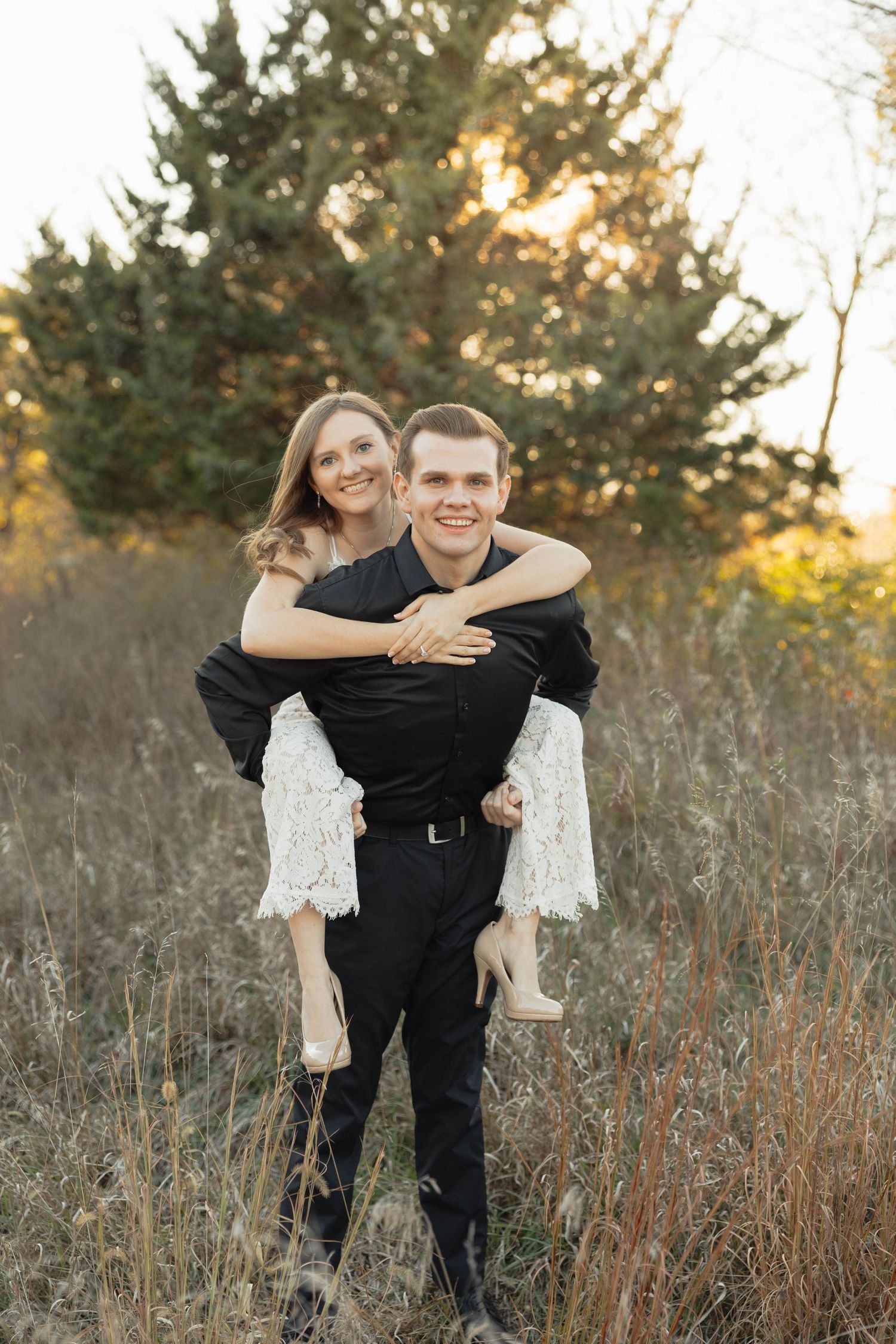 Couple embraces in a piggyback pose against tall grass and evergreen trees during golden hour sunset.