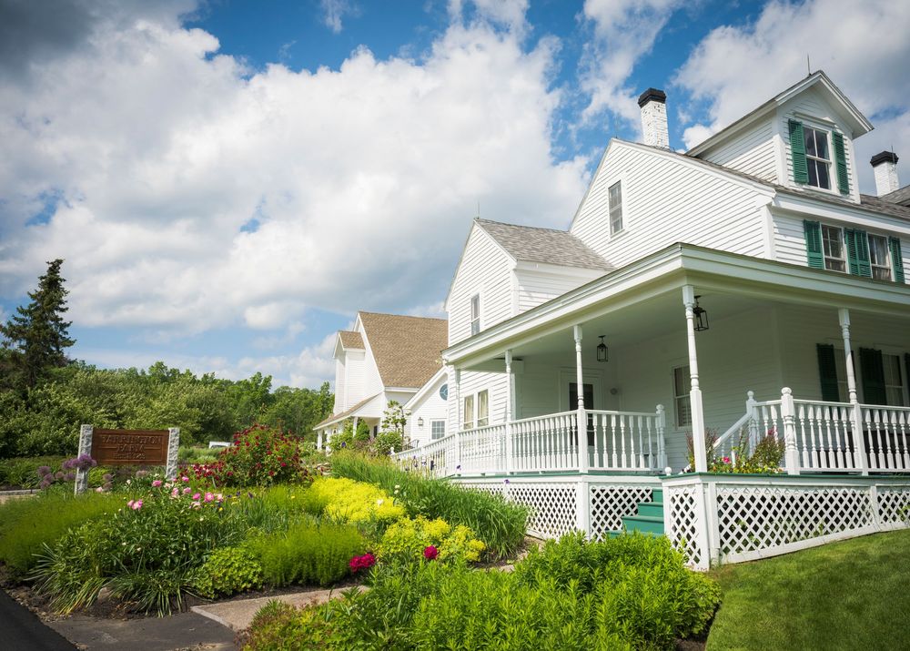 White farmhouse with wraparound porch and lattice trim surrounded by colorful flower gardens on a sunny summer day.