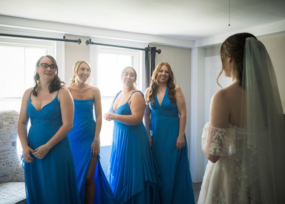 Bridesmaids in matching blue dresses gather with a bride in white gown in a sunlit room during wedding preparations.