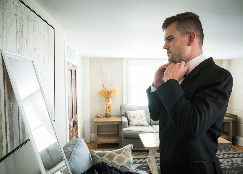 Professional in a dark suit adjusts his tie while looking in a full-length mirror in a modern living room setting.