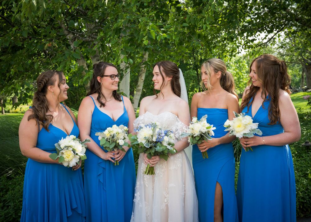A bridal party wearing bright blue bridesmaid dresses holds white flower bouquets while standing outdoors in a garden setting.