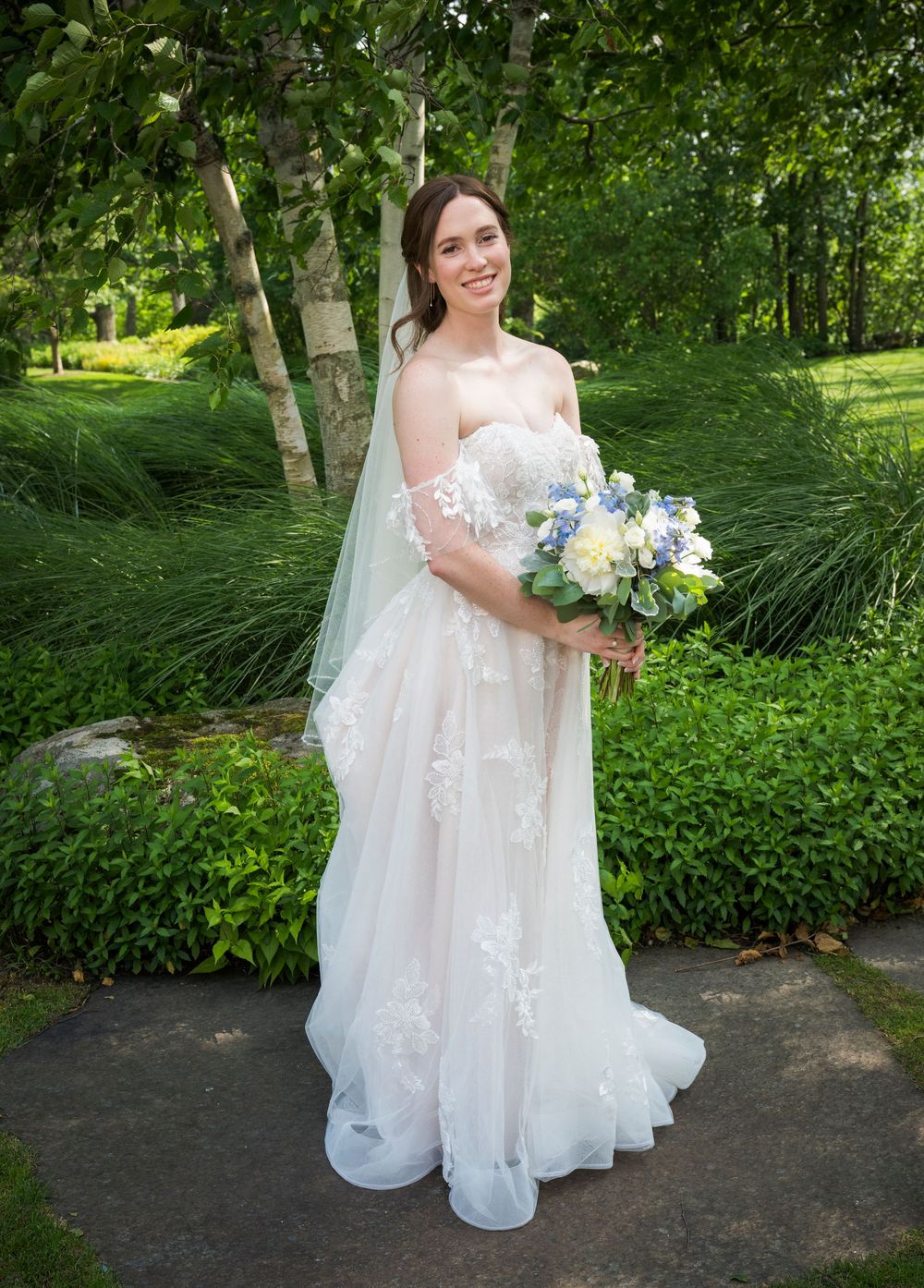 A bride in a flowing white lace gown poses in a lush garden setting holding a blue and white bouquet.