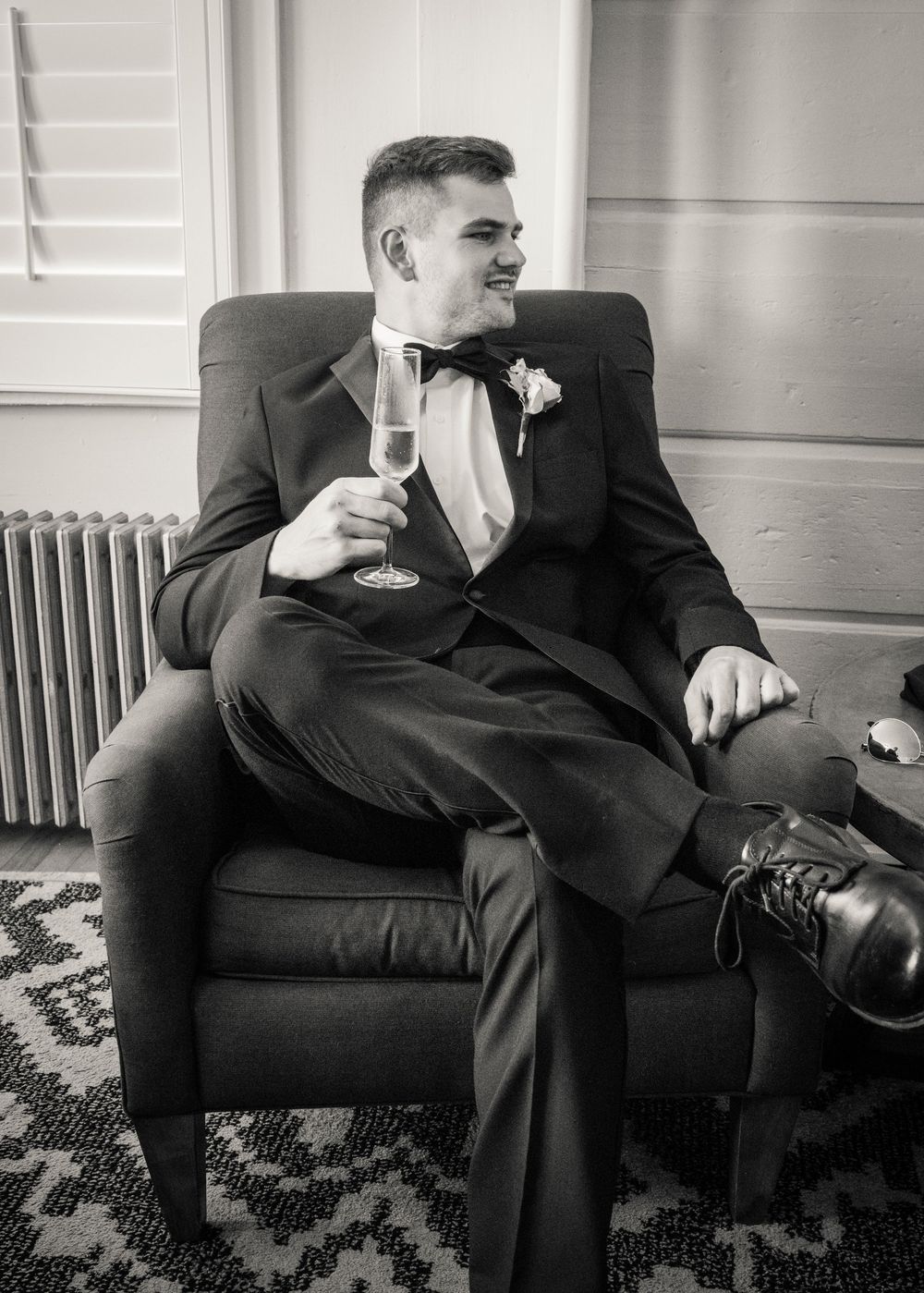 A person in a formal black tuxedo relaxes in an armchair holding a drink at a wedding celebration.