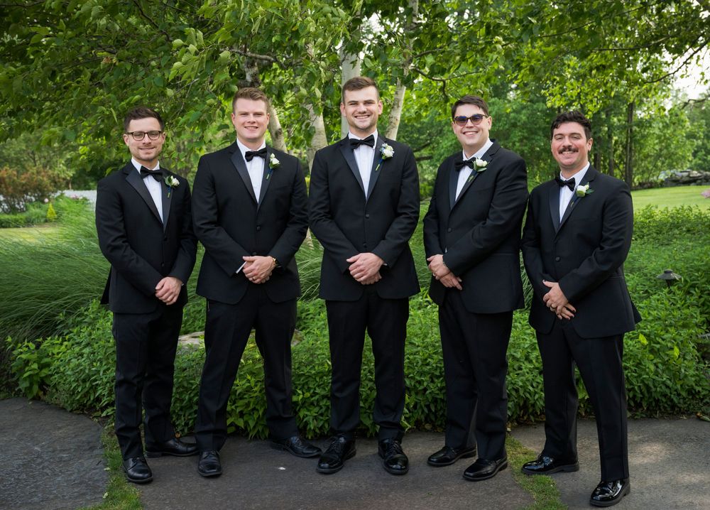 A group of groomsmen in black tuxedos pose together on a green lawn for a formal wedding photo.