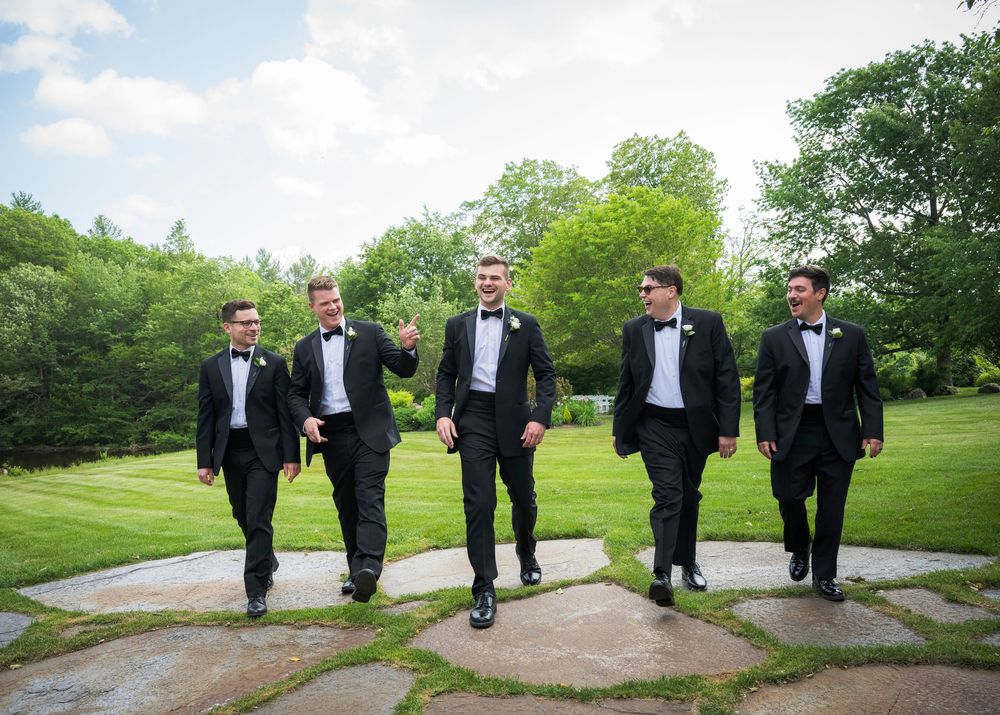 Groomsmen in black tuxedos walking in formation on a garden path.