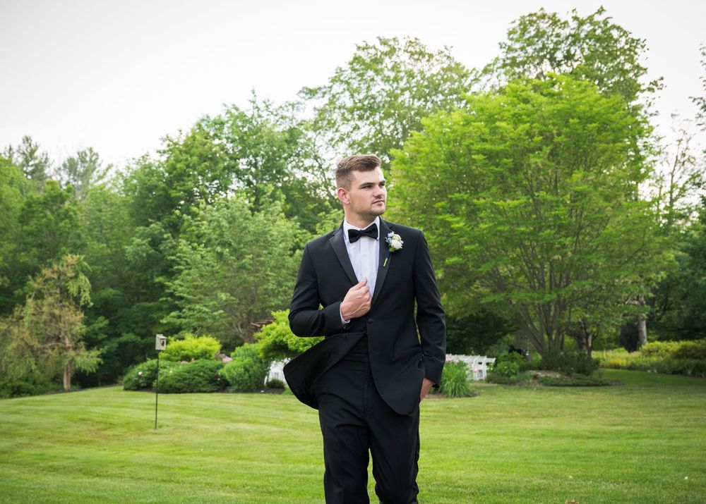 A groom in a classic black tuxedo and bowtie walks on a lush green lawn during an outdoor wedding.