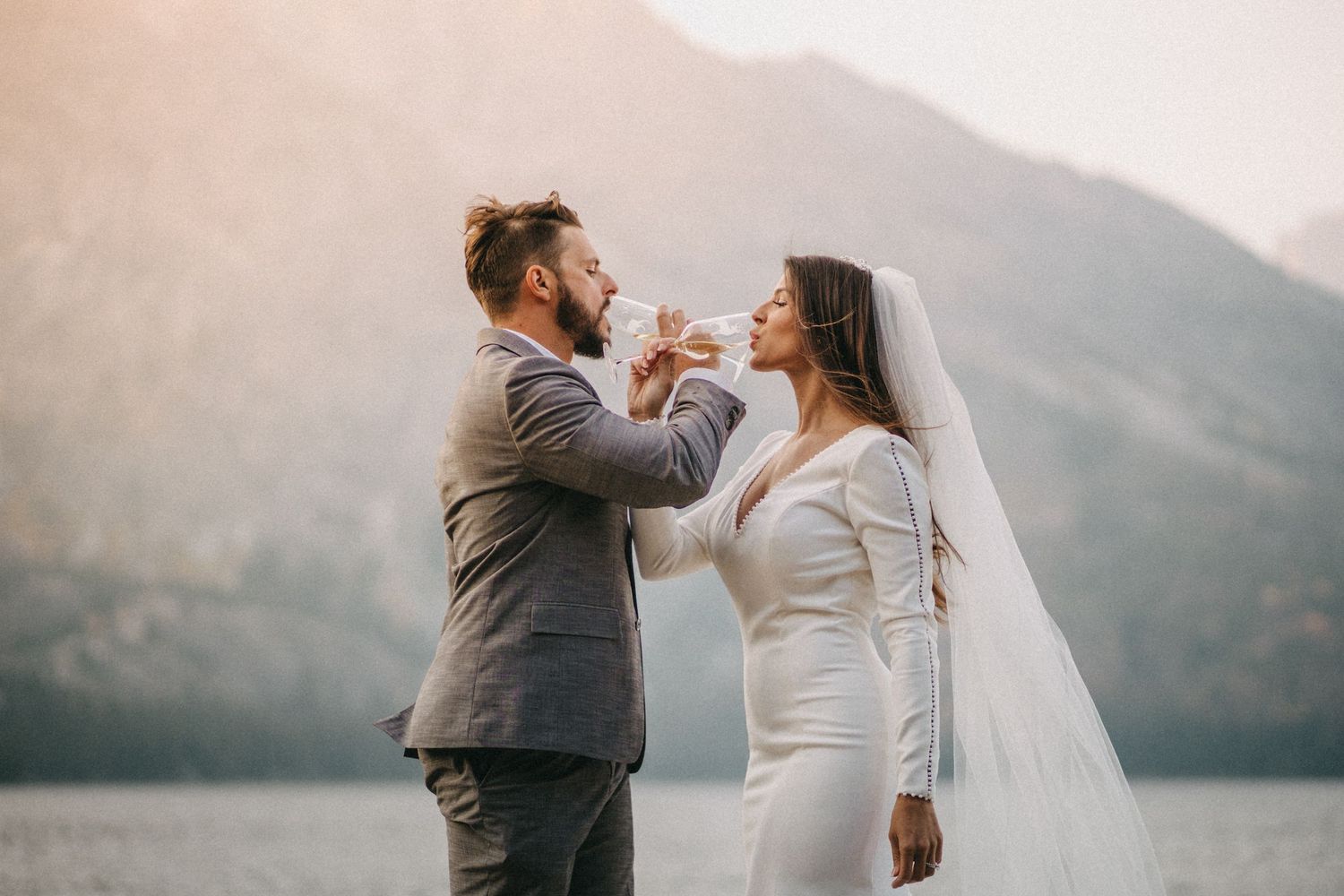 A couple in wedding attire shares a playful moment by a mountain lake at sunset.
