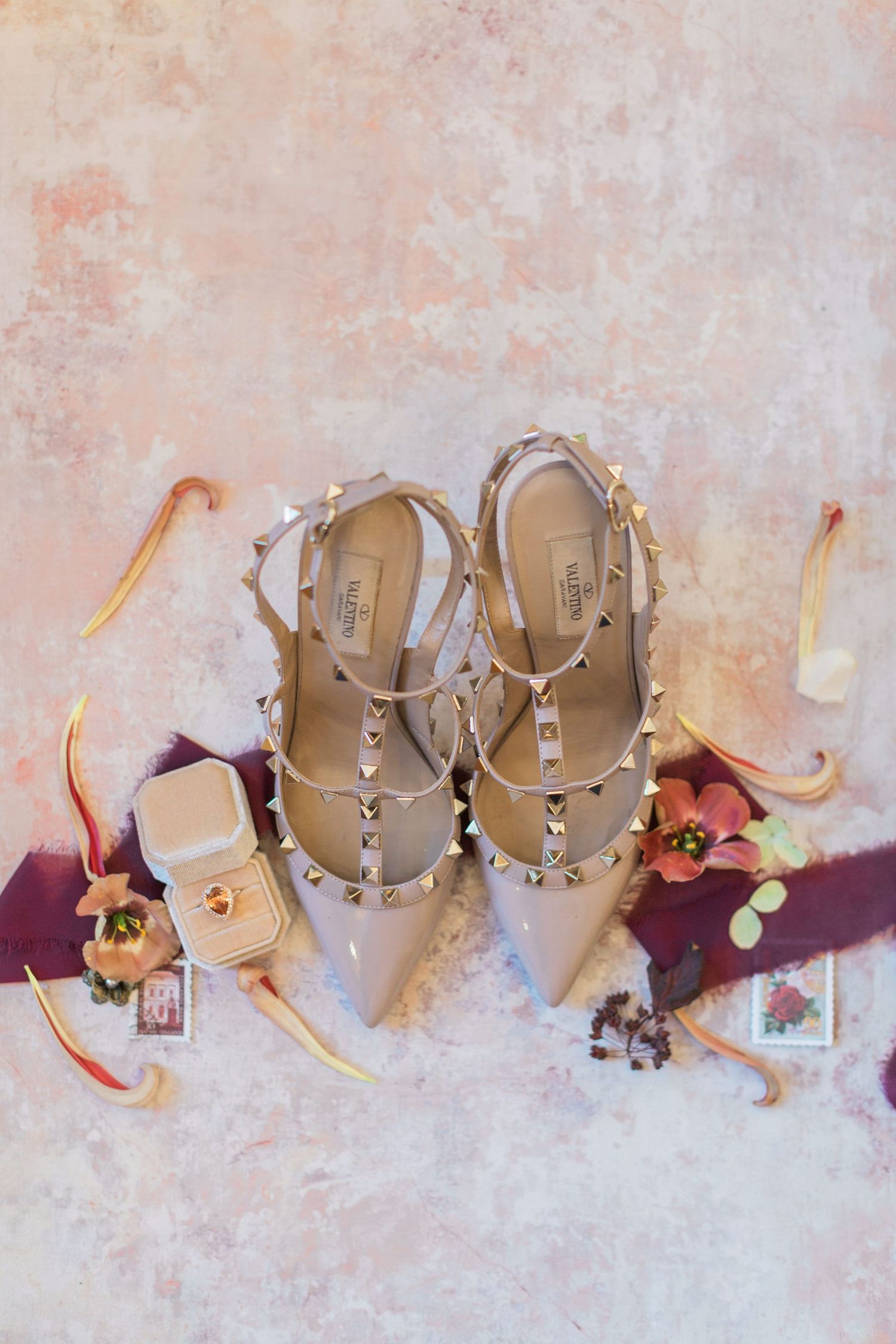 Elegant Valentino Rockstud heels with pointed toe and straps displayed with burgundy ribbon, floral flatlay and large gem ring Cotswolds