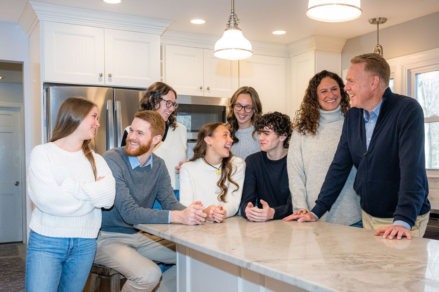 A group of people gathered around a marble kitchen island share an animated conversation in a bright modern space.