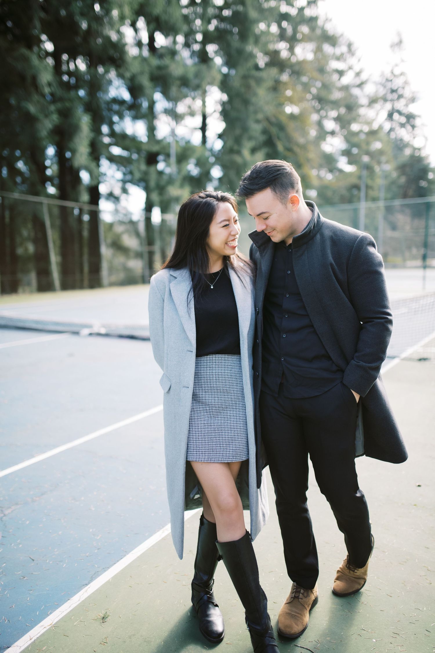 A couple in stylish winter coats sharing a romantic moment while walking on a tennis court.