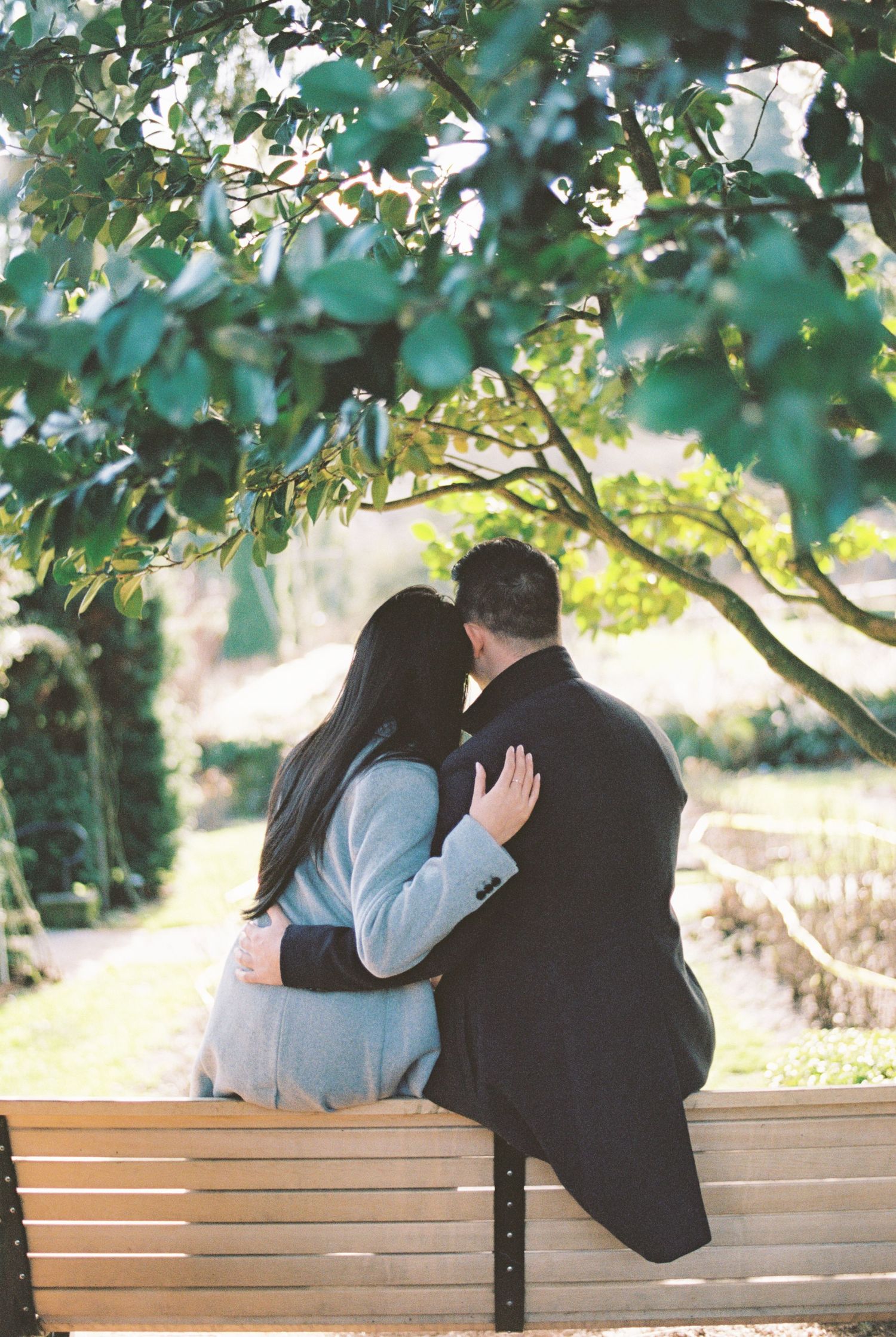 A couple shares tender moments while sitting together on a wooden park bench under green foliage.