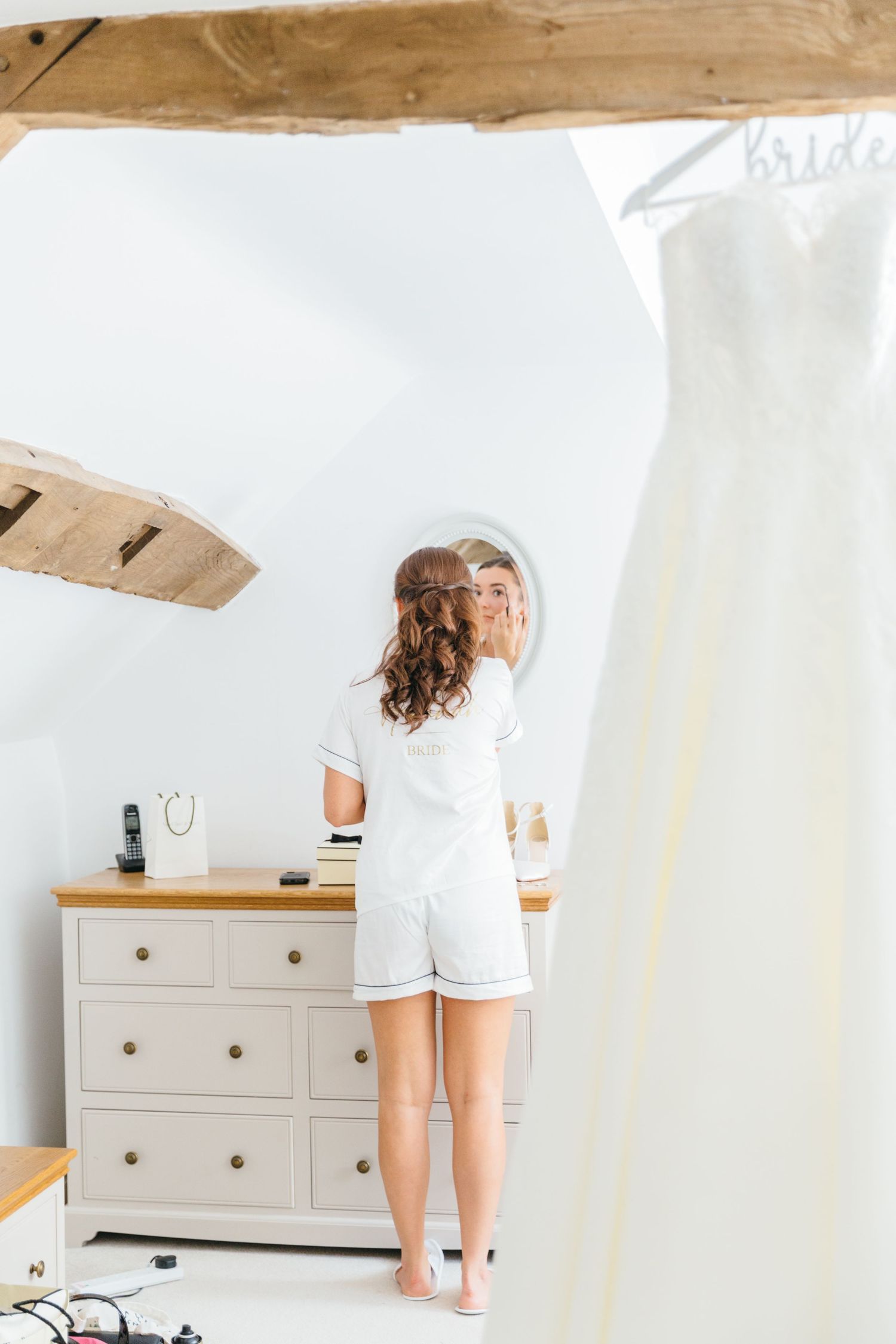 A bride to be in white PJs stands in a bright minimalist bedroom with wooden beams with wedding dress hanging. Lapstone Barn wedding