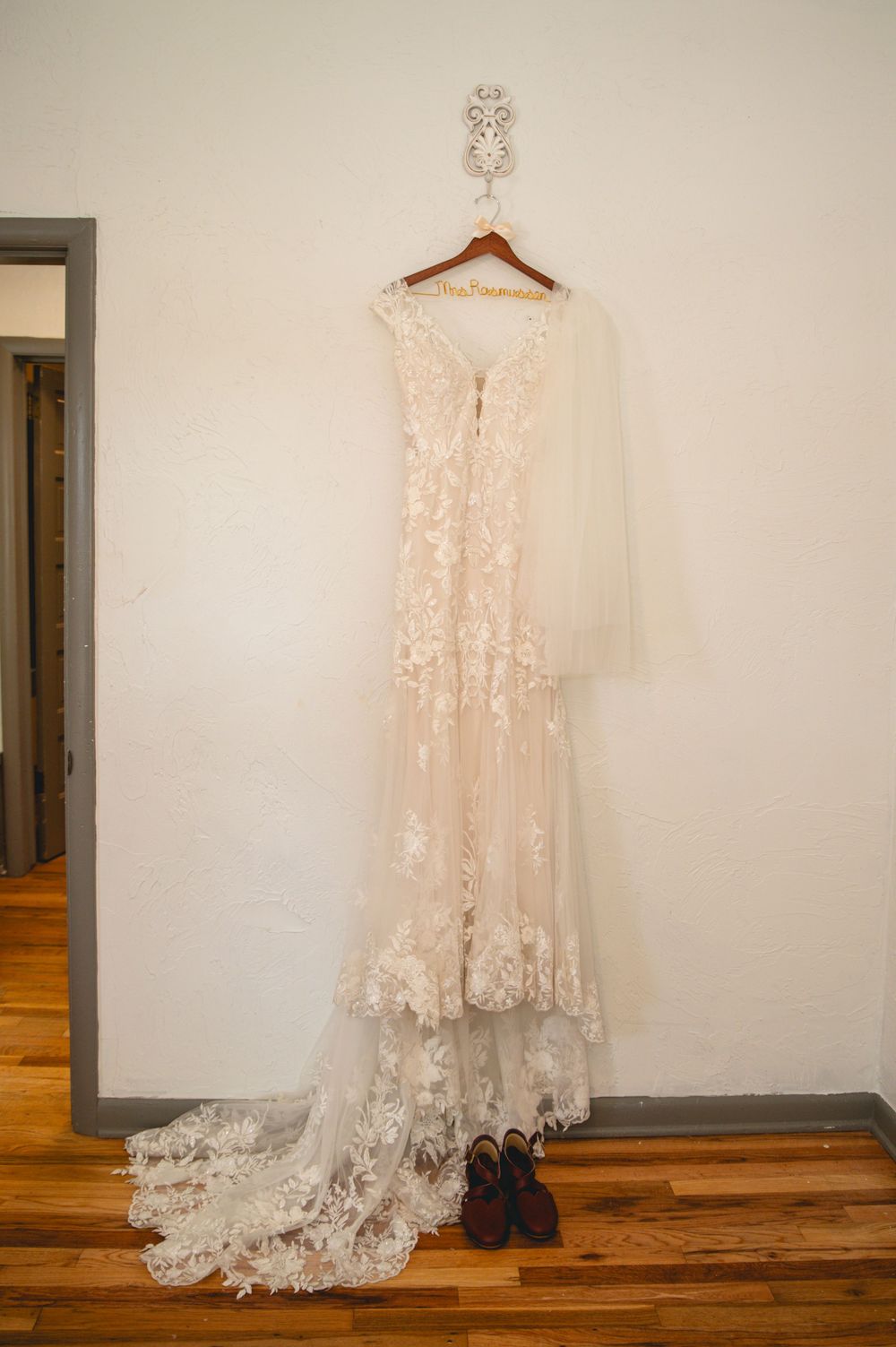 Elegant lace wedding dress with long train hanging on wooden hanger against white wall with black boots on hardwood floor.