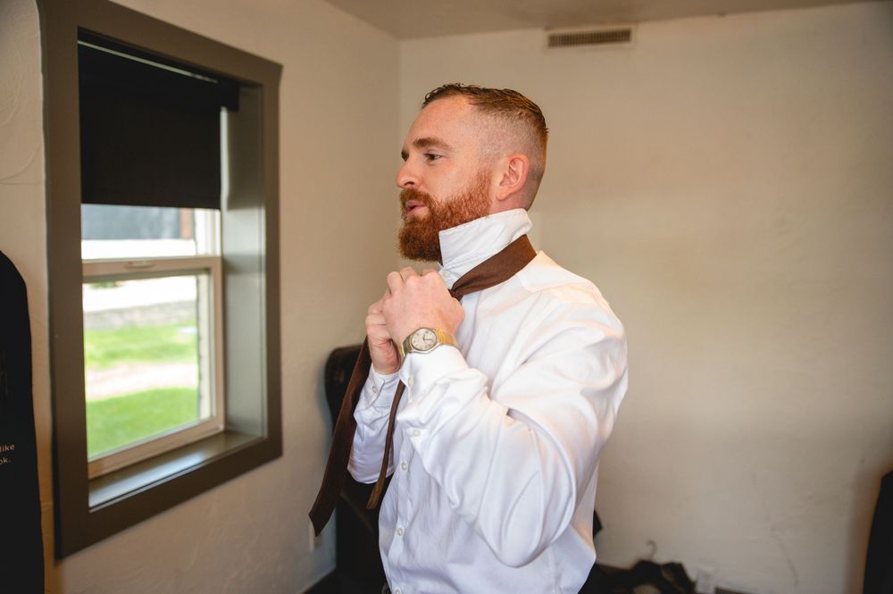 A person in a white dress shirt adjusts their tie while standing near a window in a small room.