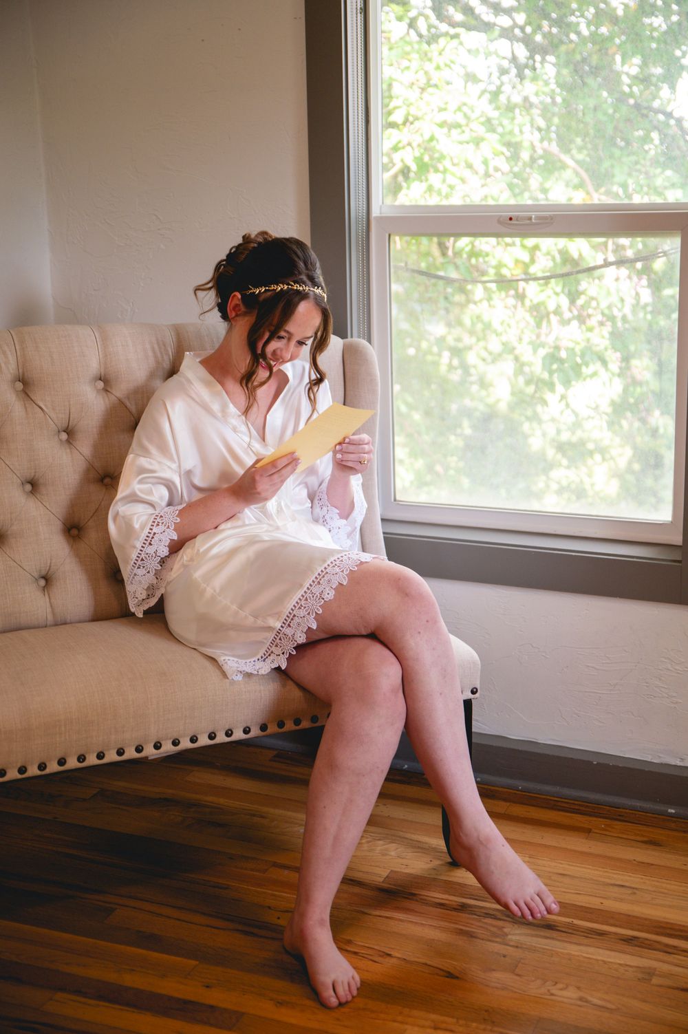 Person in white lace dress sitting on tufted beige couch by window with natural light on hardwood floors.